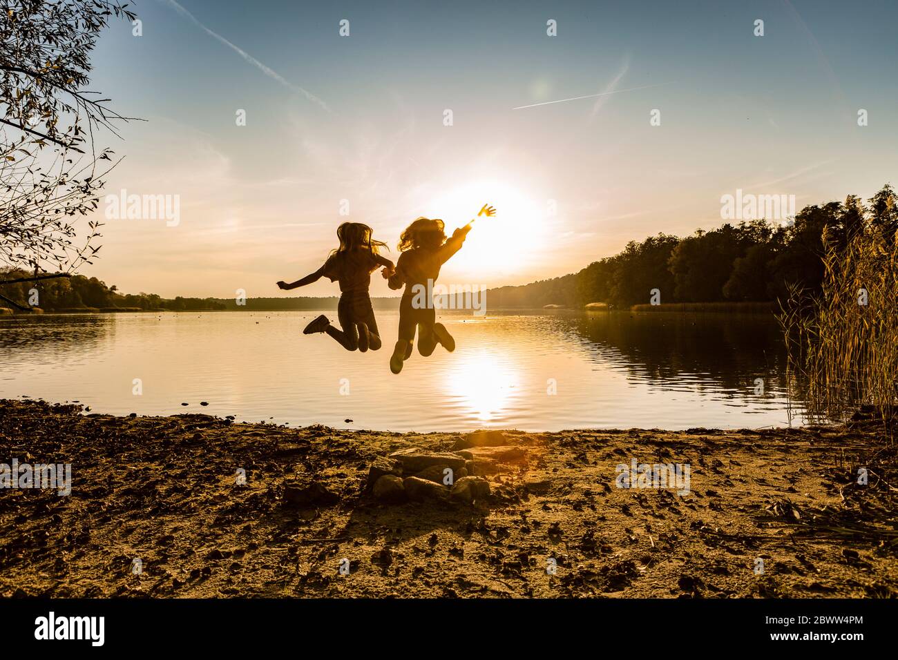Freunde springen in der Luft, während sie am Seeufer während des Sonnenuntergangs genießen Stockfoto