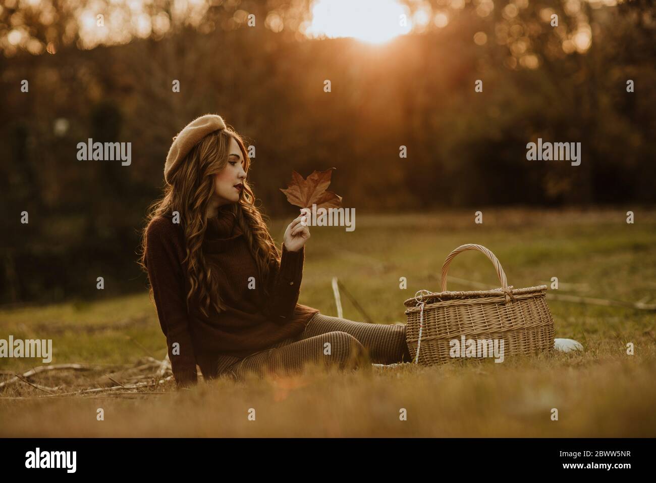 Modische Frau mit Korb und Herbstblatt sitzt auf einer Wiese bei Hintergrundbeleuchtung Stockfoto