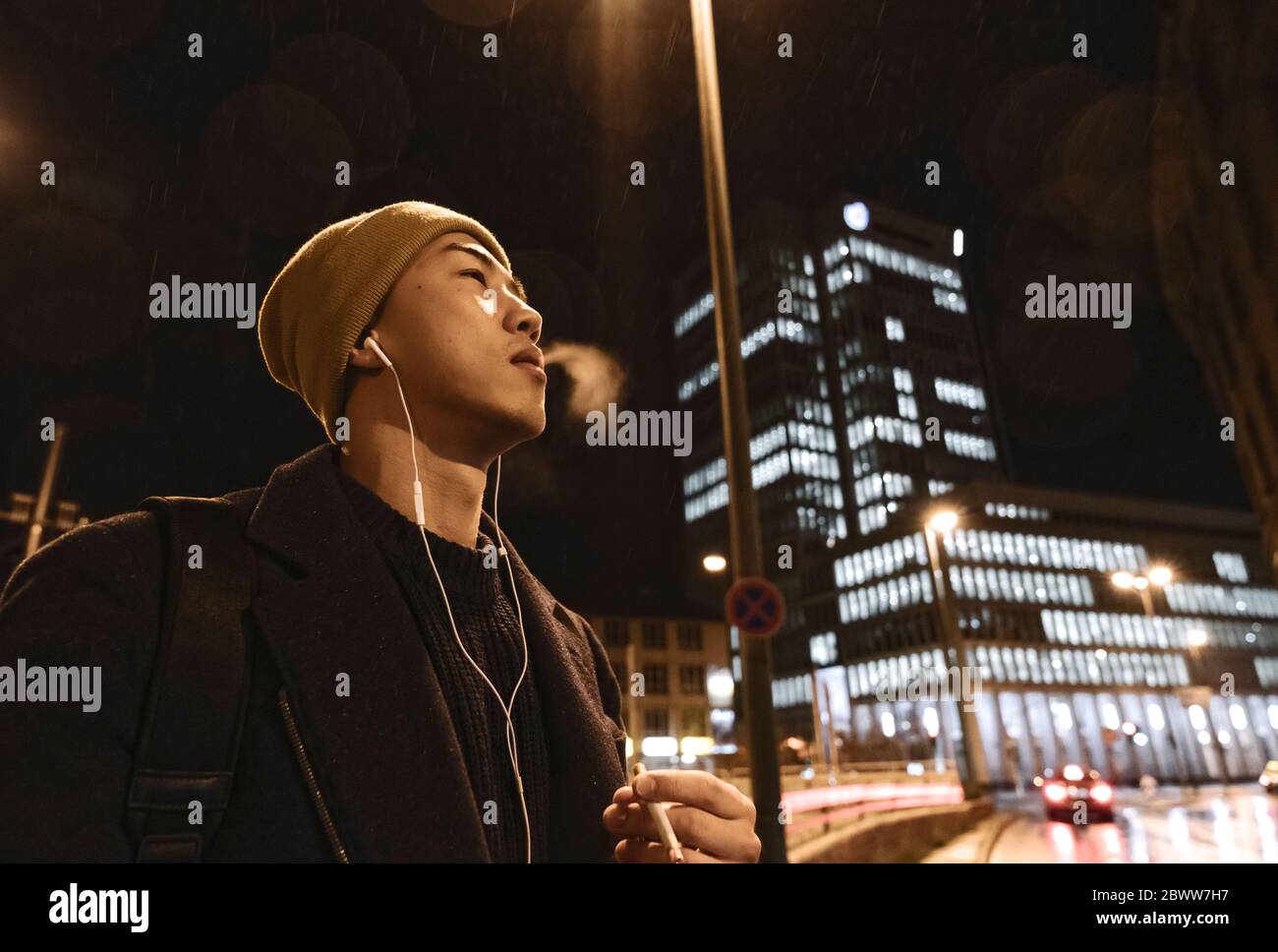 Stilvoller Mann mit gelbem Hut und Ohrhörer, der nachts in der Stadt eine Zigarette raucht Stockfoto