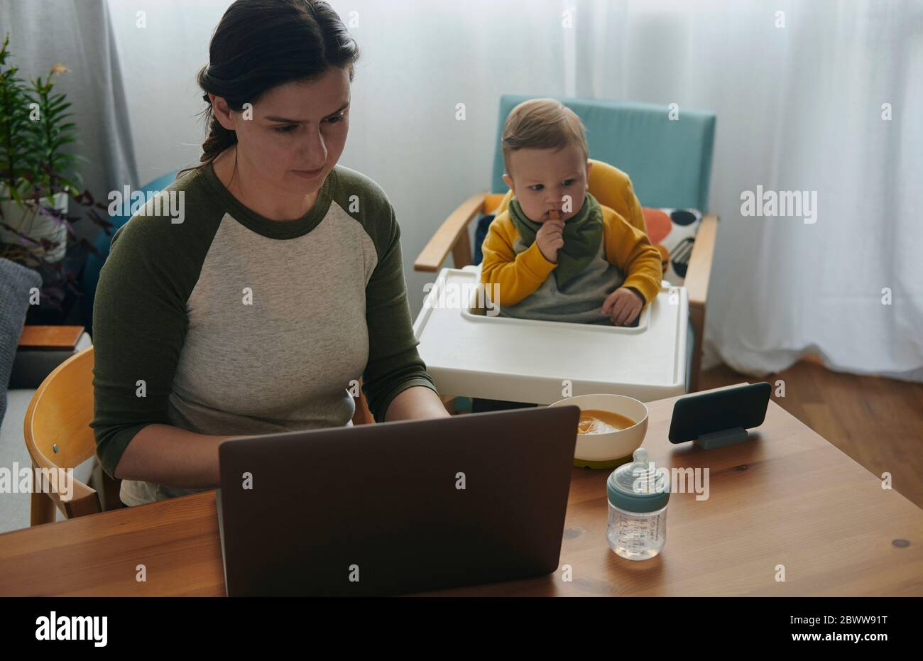 Frau, die zu Hause am Laptop arbeitet, während ihr kleiner Sohn Filme auf dem Handy sieht Stockfoto