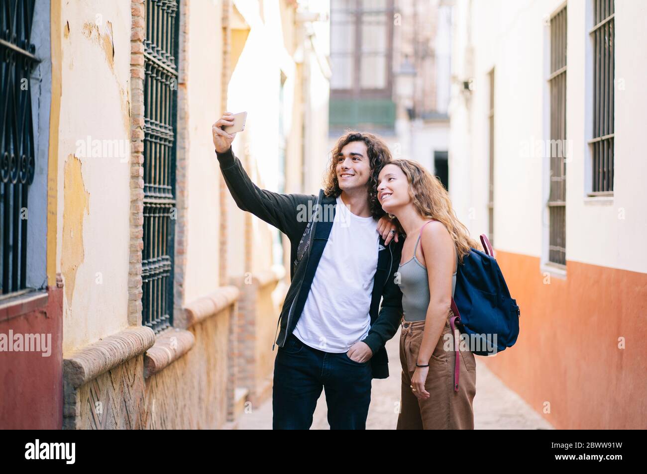 Lächelndes junges Paar, das Selfie macht, während es in der engen Straße in Santa Cruz, Sevilla, Spanien steht Stockfoto