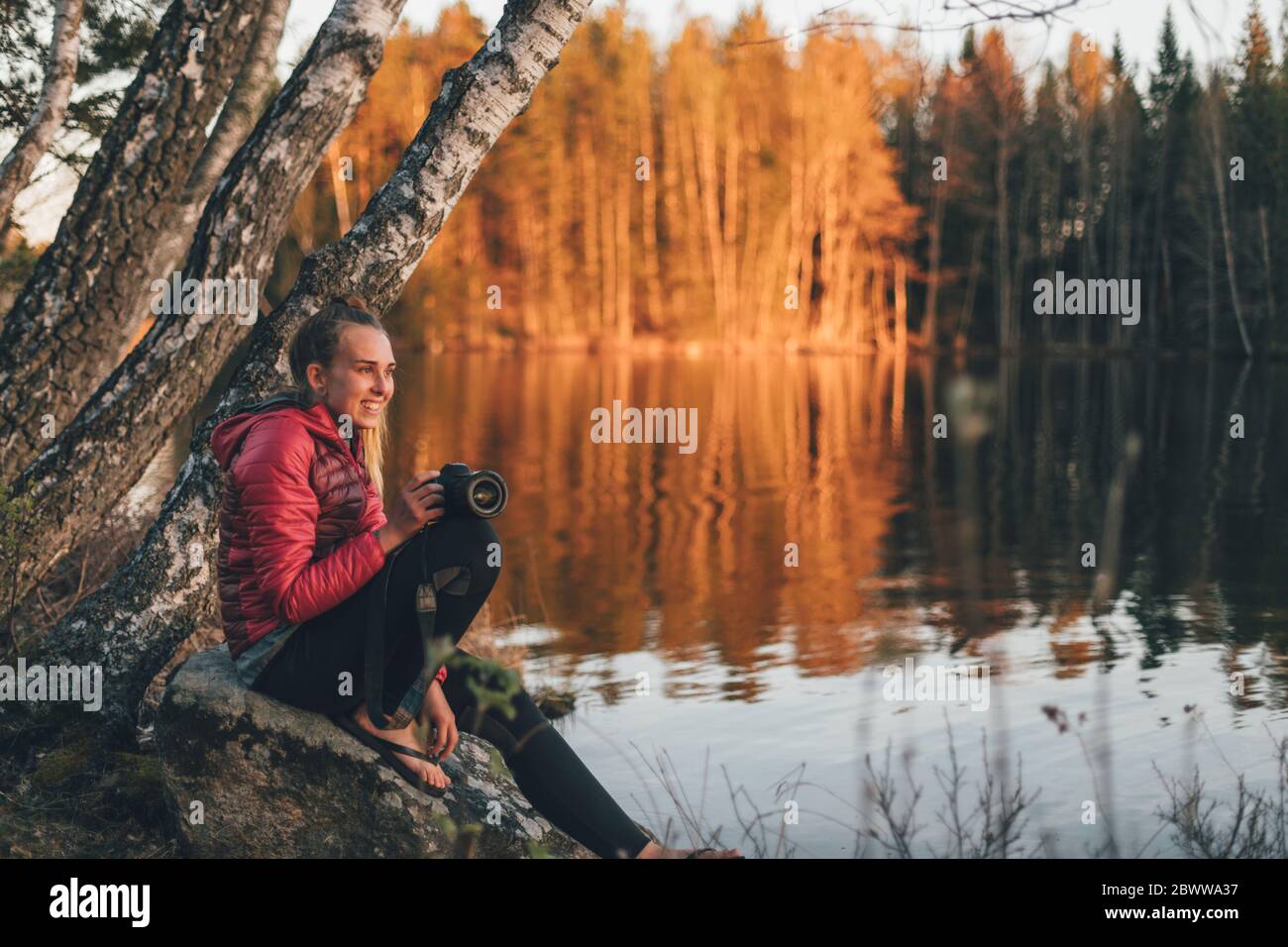 Junge Frau, die auf Stein am Seeufer sitzt und den Sonnenuntergang betrachtet Stockfoto