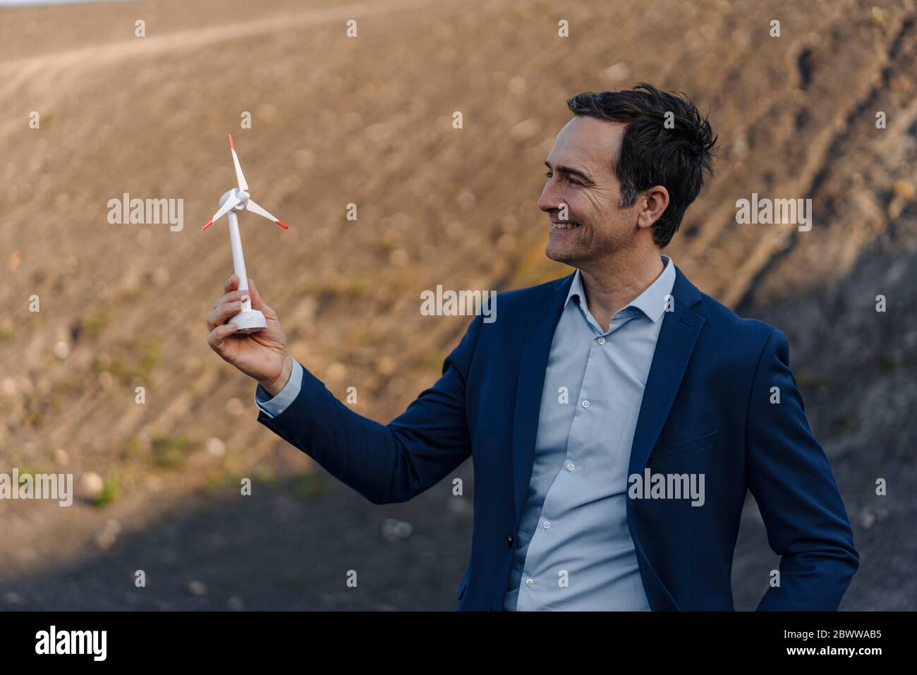 Glücklicher reifer Geschäftsmann auf einem stillgehaltenem Minenspitze, der Windturbinenmodell hält Stockfoto