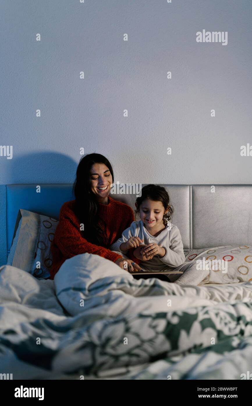 Lachende Mutter und ihre kleine Tochter sitzen zusammen auf dem Bett und schauen sich Bilderbuch an Stockfoto