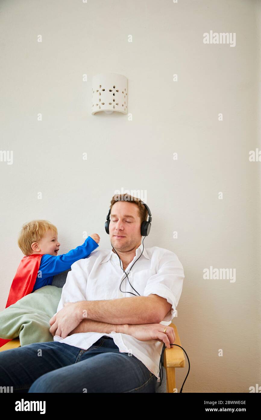 Junge in Superman-Kostüm und nerviger Vater, während er mit Kopfhörern Musik spielt Stockfoto