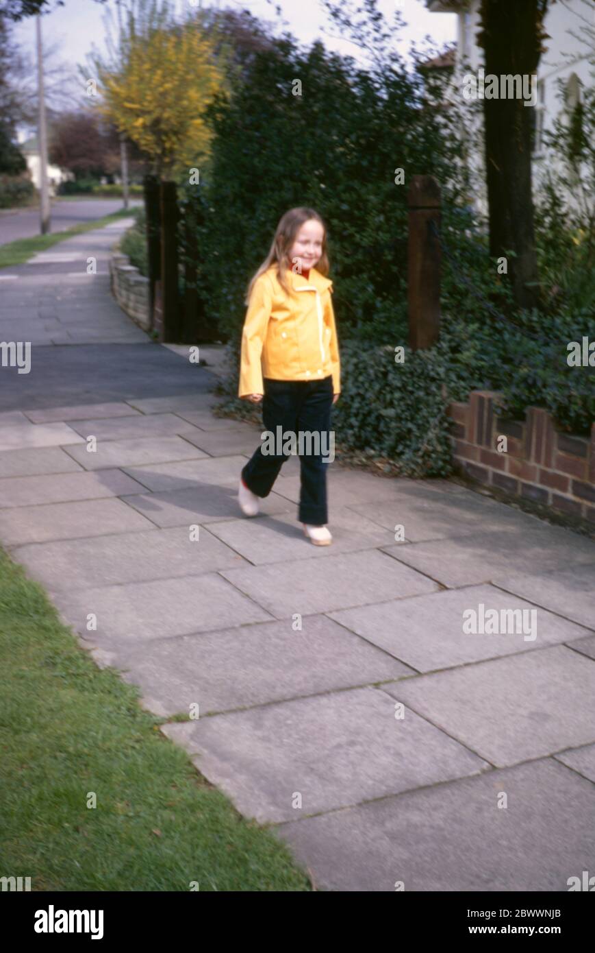 Ein junges, 7-jähriges Mädchen mit langen, schönen Haaren, das eine gelbe Jacke, cremefarbene Clogs und dunkelblaue Schnüre trägt, geht vor ihrem Familienhaus in den Londoner Vororten Hatch End, Middlesex, Großbritannien, auf dem Bürgersteig entlang Stockfoto