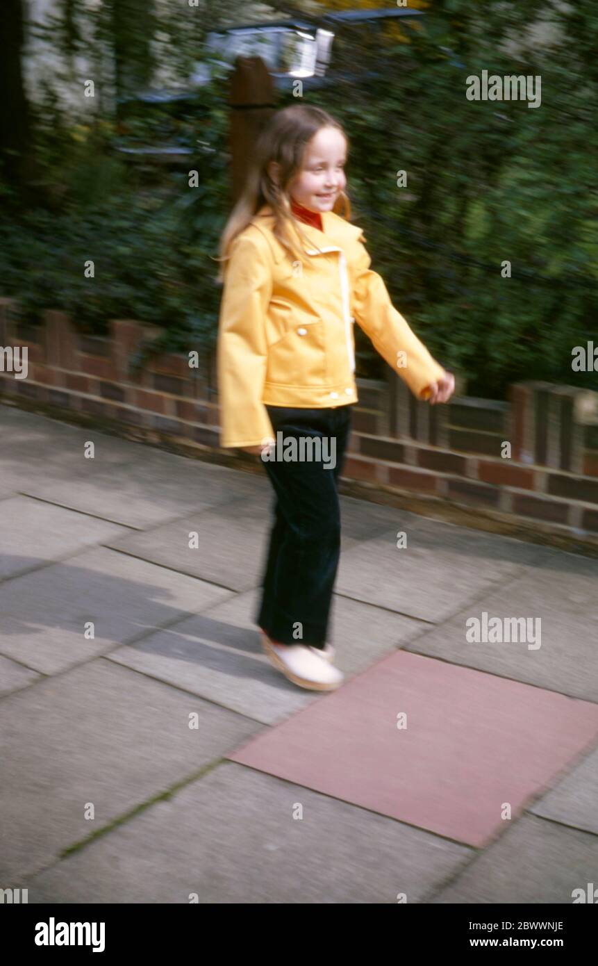 Ein junges, 7-jähriges Mädchen mit langen, schönen Haaren, das eine gelbe Jacke, cremefarbene Clogs und dunkelblaue Schnüre trägt, geht vor ihrem Familienhaus in den Londoner Vororten Hatch End, Middlesex, Großbritannien, auf dem Bürgersteig entlang Stockfoto