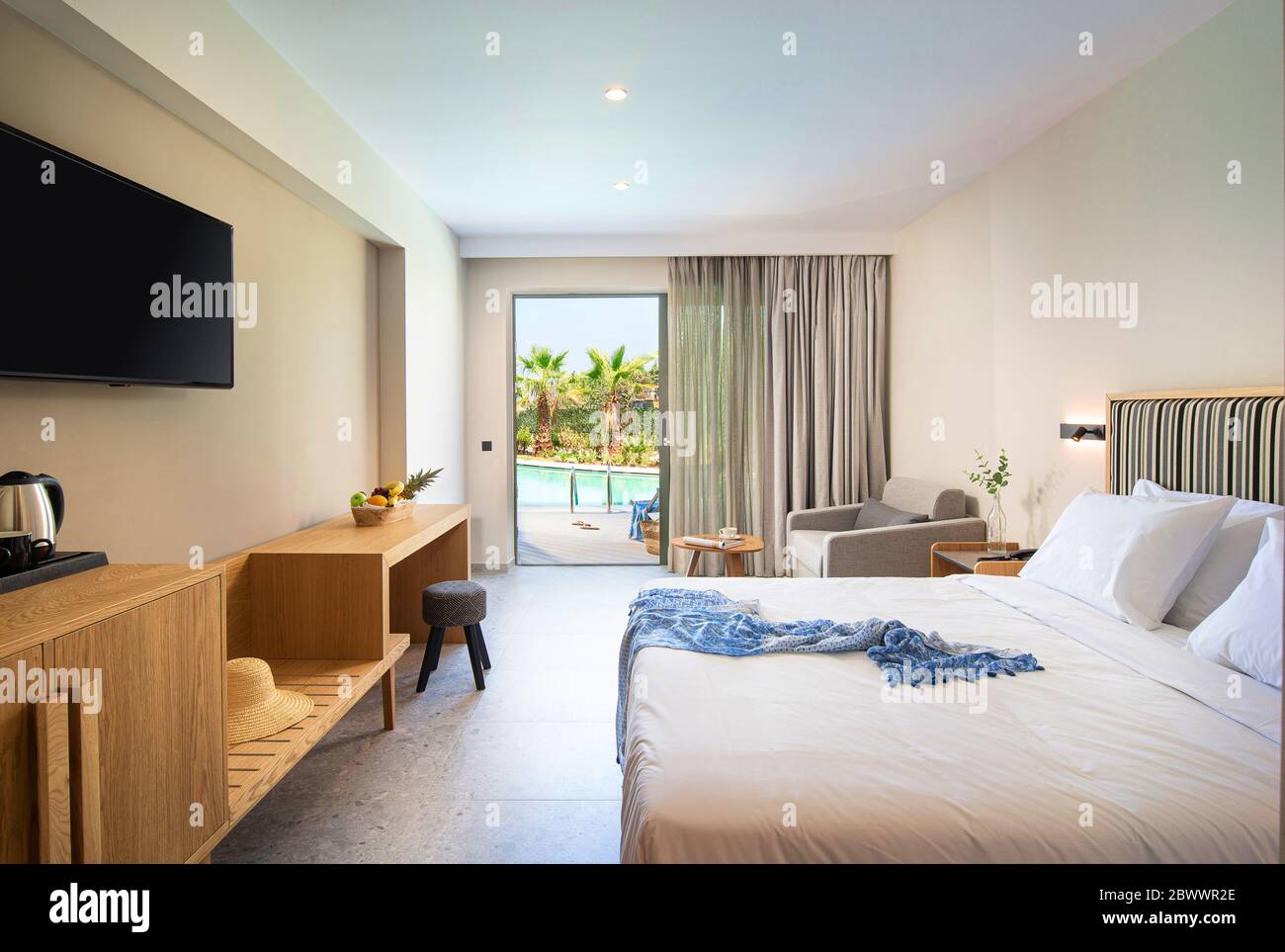 Beige gestreifte und helle Holzeinrichtung in modernem minimalistischem Stil Schlafzimmer mit offenem französischem Fenster und Blick auf den tropischen Swimmingpool Stockfoto