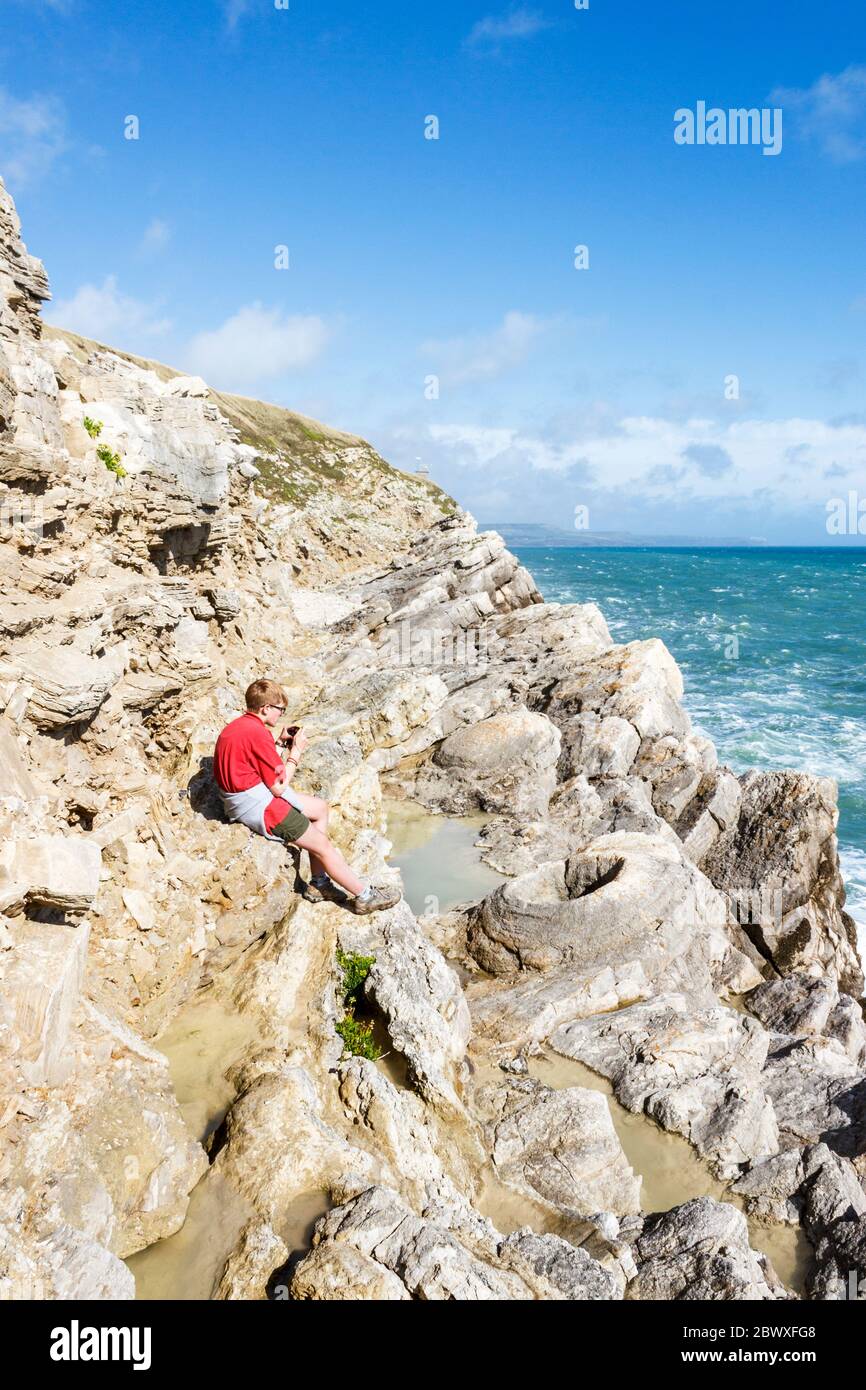 Ein 15-jähriges Mädchen in einem roten Hemd, das auf Felsen sitzt und im fossilen Wald an der Küste bei Lulworth, Dorset, Großbritannien, fotografiert Stockfoto