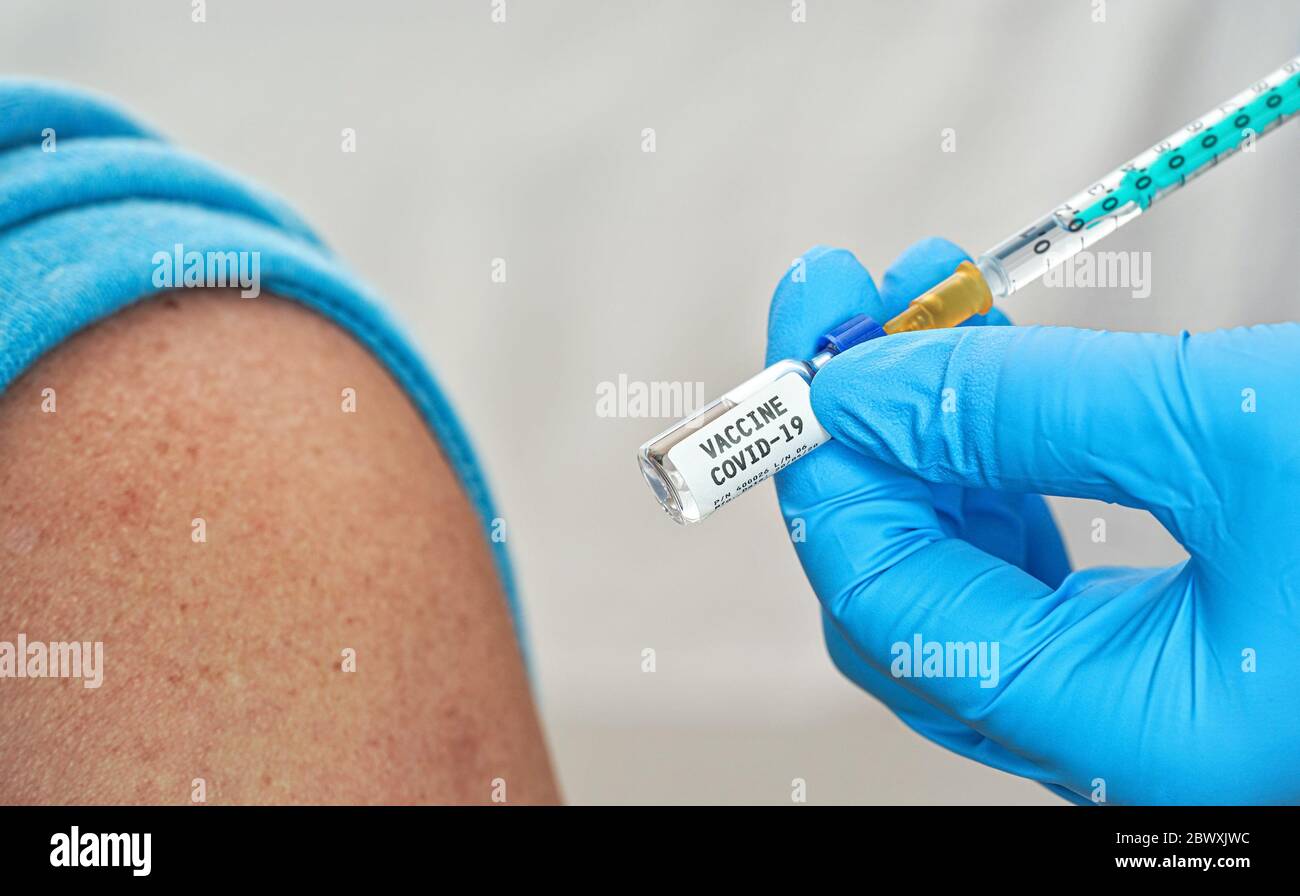 Hand in blauen Handschuhen hält Spritze und Coronavirus-Impfstoff (Aufkleber eigenes Design), über es auf Schulter des Patienten in der Nähe sitzen injizieren, Nahaufnahme deta Stockfoto