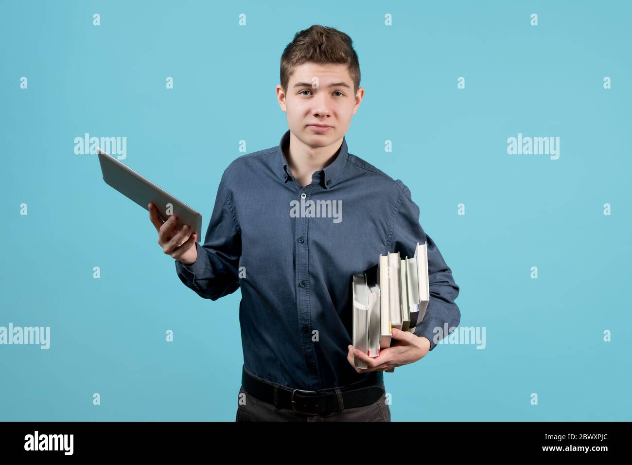 Ein Stapel Bücher unter der Achselhöhle eines Teenagers und ein Tablet in der anderen Hand. Ein Teenager schaut mit einem Lächeln, zeigt die Unannehmlichkeiten eines Stapels Stockfoto