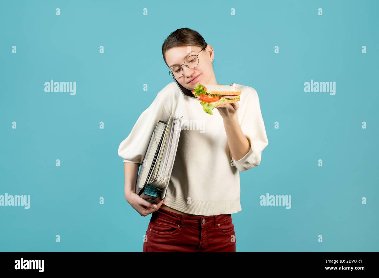 Ein Mädchen mit einer Brille mit Dokumenten unter der Achselhöhle telefoniert und ist dabei, ein Sandwich zu essen. Mit einem Lächeln auf ein Sandwich schauen Stockfoto