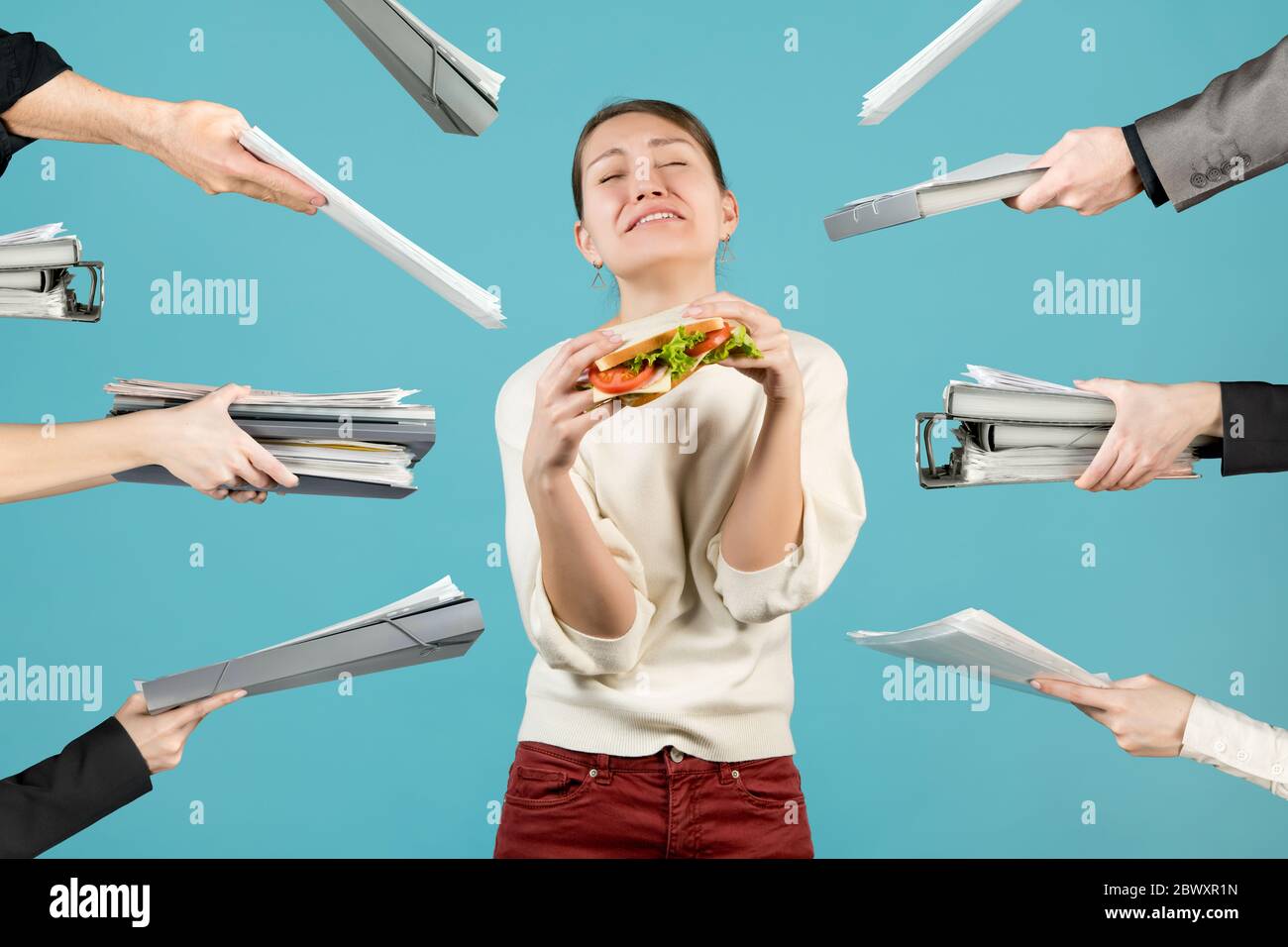 Eine junge Frau leidet unter der Unfähigkeit, ein Sandwich zu essen, das sie in den Händen hält, weil sie viel Arbeit hat. Konzept der Überlastung Stockfoto