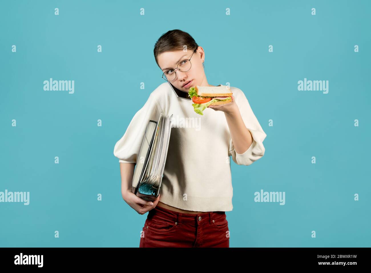 Ein Mädchen mit einer Brille mit Dokumenten unter der Achselhöhle telefoniert und ist dabei, ein Sandwich zu essen. Schnelles Snack-Konzept, Essen für unterwegs. Stockfoto