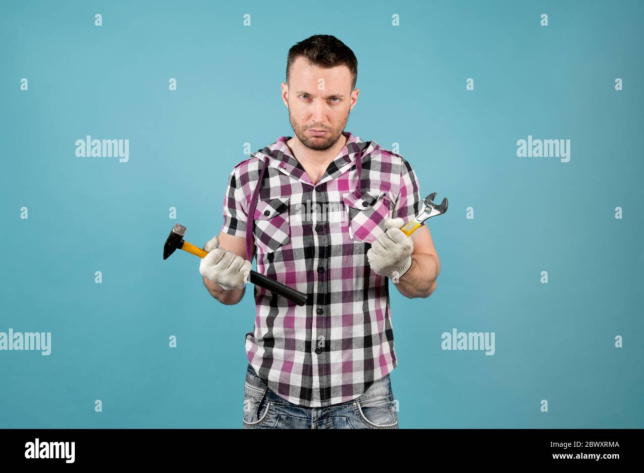 Unzufrieden harter Arbeiter mit einem Hammer und verstellbarem Schraubenschlüssel in den Händen, der auf die Kamera schaut. Sein Gesicht und seine Hände sind schmutzig und tragen Schutzhandschuhe Stockfoto