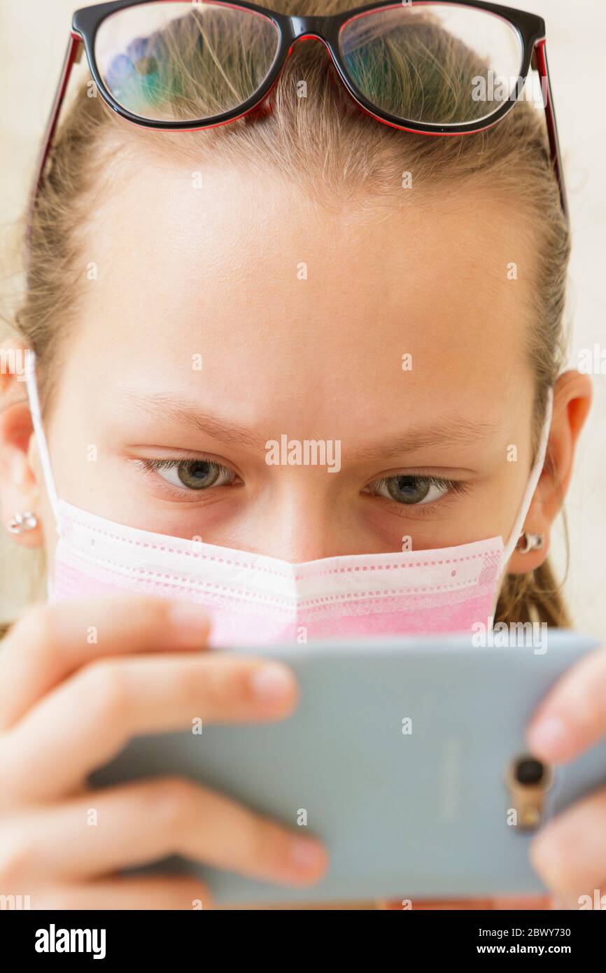 Mädchen in einer medizinischen Maske mit Sehstörungen und Brille auf dem Kopf schaut auf das Telefon. Stockfoto