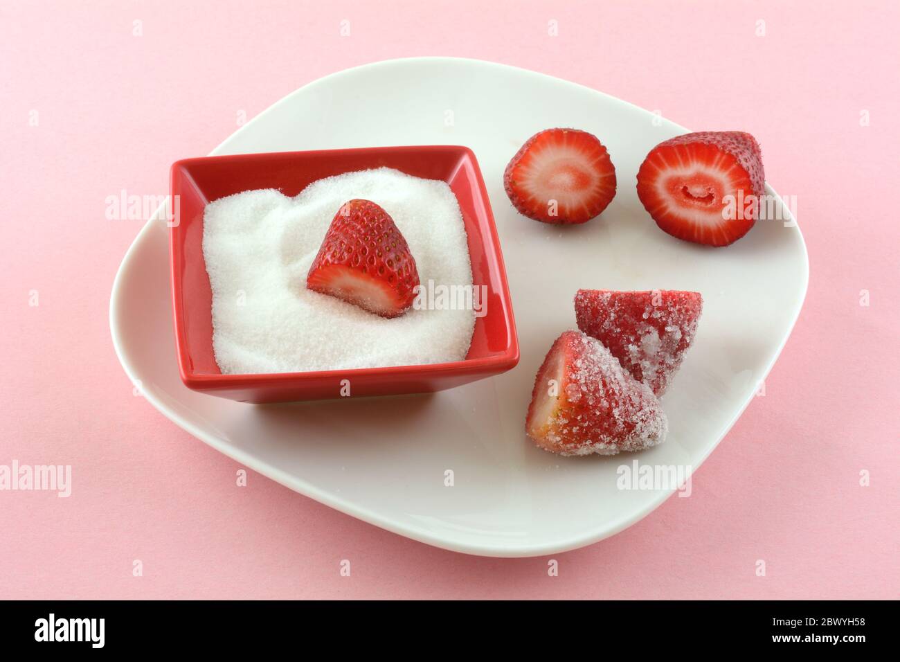 Frische roh geschnittene Erdbeeren und Zucker in roter Zuckerdose auf weißem Teller auf rosa Hintergrund Stockfoto