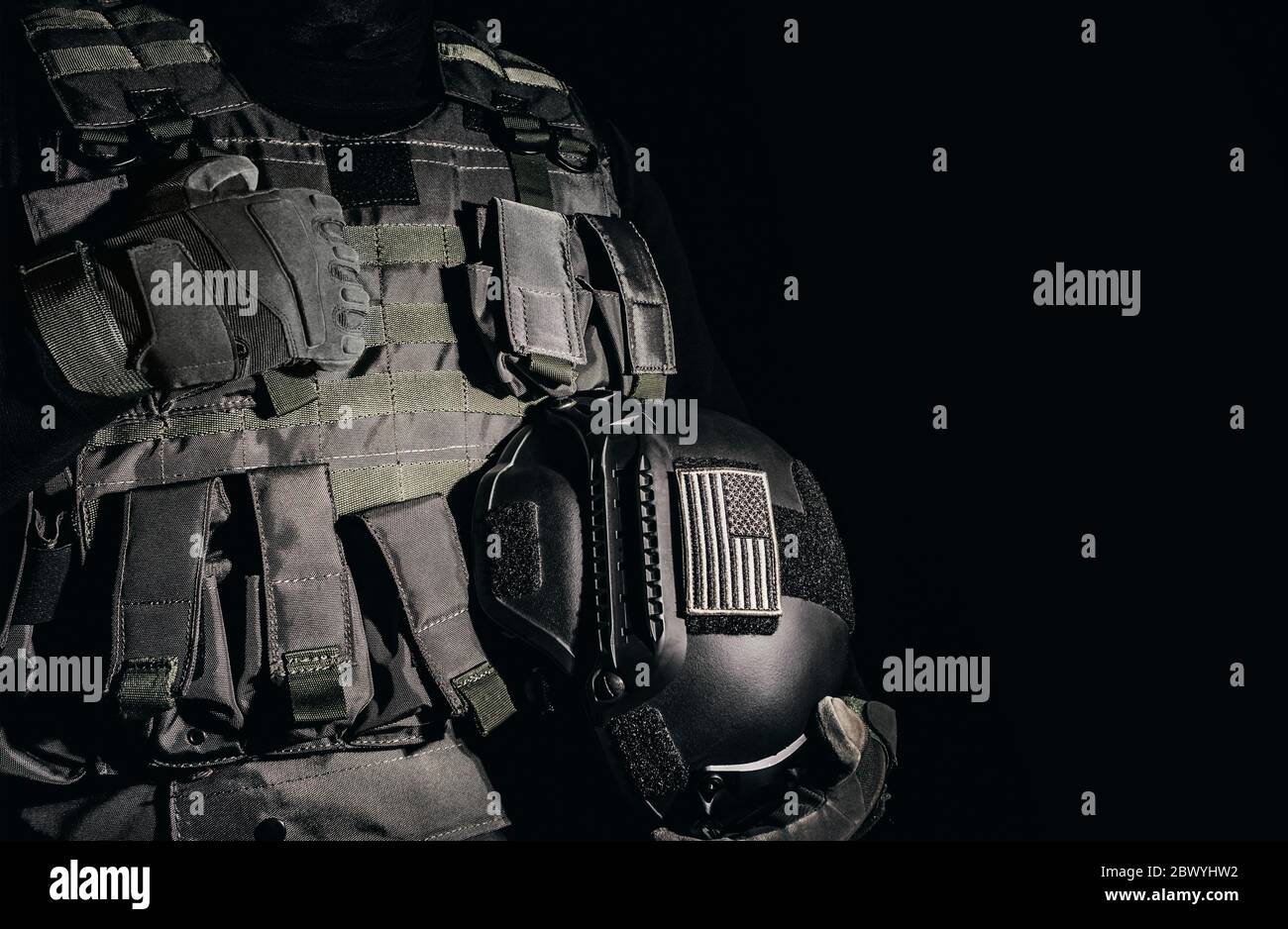 Foto eines Soldaten in taktischen Handschuhen und Rüstungsweste stehend und hält Militärhelm mit amerikanischen Flagge Patch auf schwarzem Hintergrund. Stockfoto