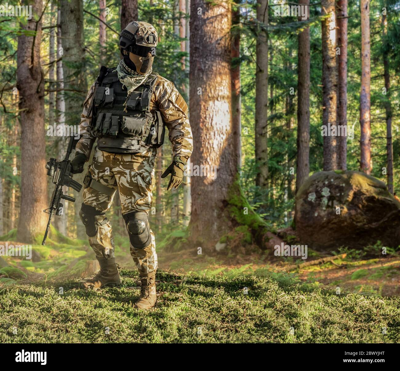Foto eines voll ausgestatteten Soldaten in Outfit, Rüstung, Helm, Brille mit automatischem Gewehr posiert auf Waldhintergrund. Stockfoto