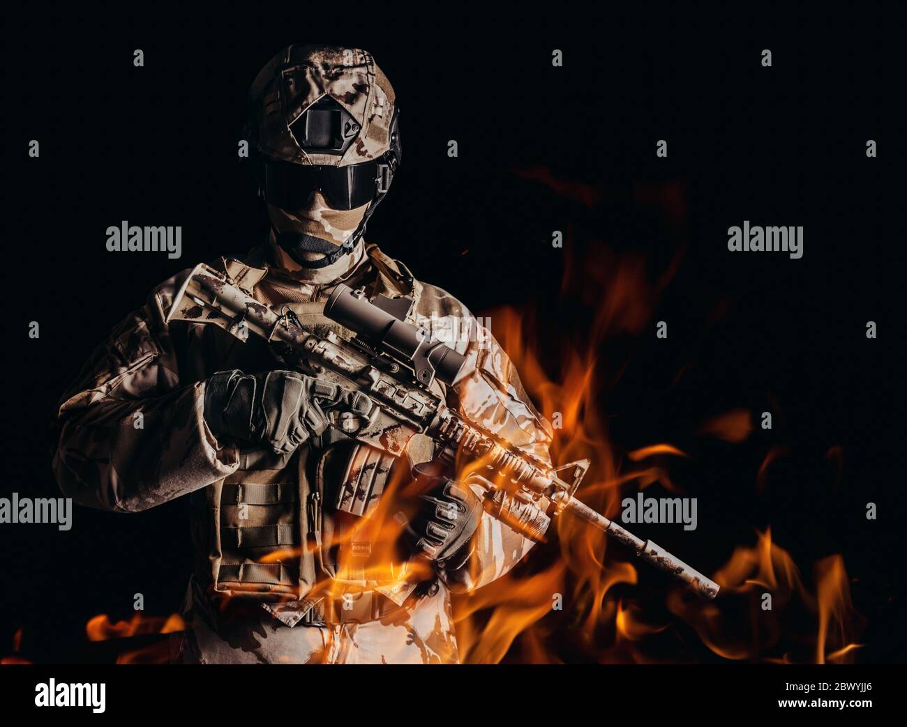 Foto eines voll ausgestatteten Wüstensoldaten in getarnter Rüstung, Handschuhe und Helm, der das automatische Gewehr in Feuer auf schwarzem Hintergrund hält. Stockfoto
