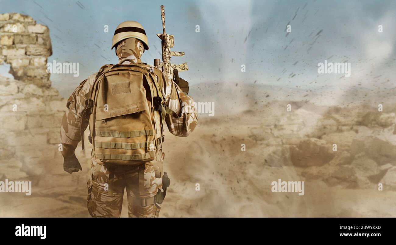 Foto eines voll ausgestatteten Soldaten in Uniform, Rüstung, Helm, Rucksack und Gewehr stehend Rückansicht auf Wüste Ruinen Hintergrund mit Staub fliegen. Stockfoto