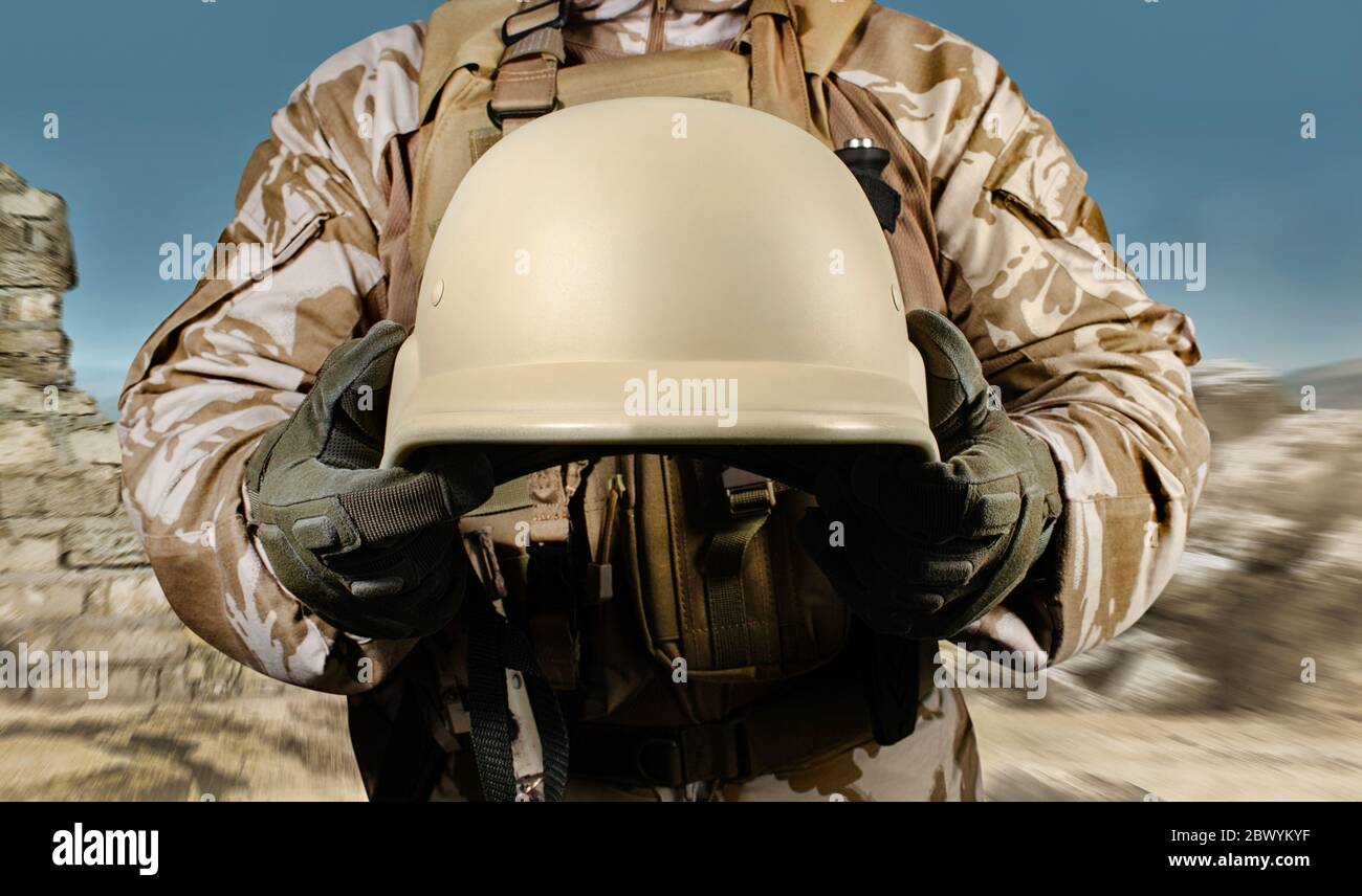 Foto eines voll ausgestatteten Soldaten in Uniform, Rüstung hält Helm in Wüste Schlachtfeld Hintergrund Nahaufnahme Vorderansicht. Stockfoto