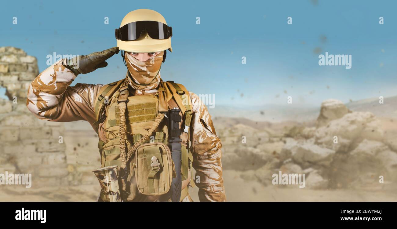 Foto eines voll ausgestatteten Soldaten in Uniform, Rüstung, Helm und Brille stehend salutieren in Wüste Schlachtfeld Hintergrund. Stockfoto