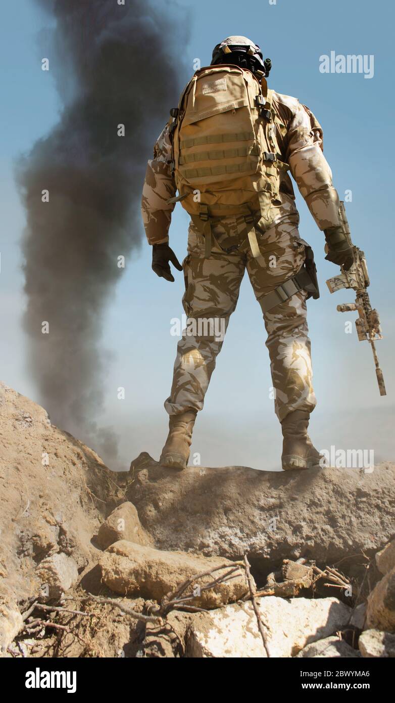 Foto eines voll ausgestatteten Wüstensoldaten mit Gewehr auf Steinberg mit  taktischen Rucksack und Rauch auf Hintergrund Rückansicht stehen  Stockfotografie - Alamy