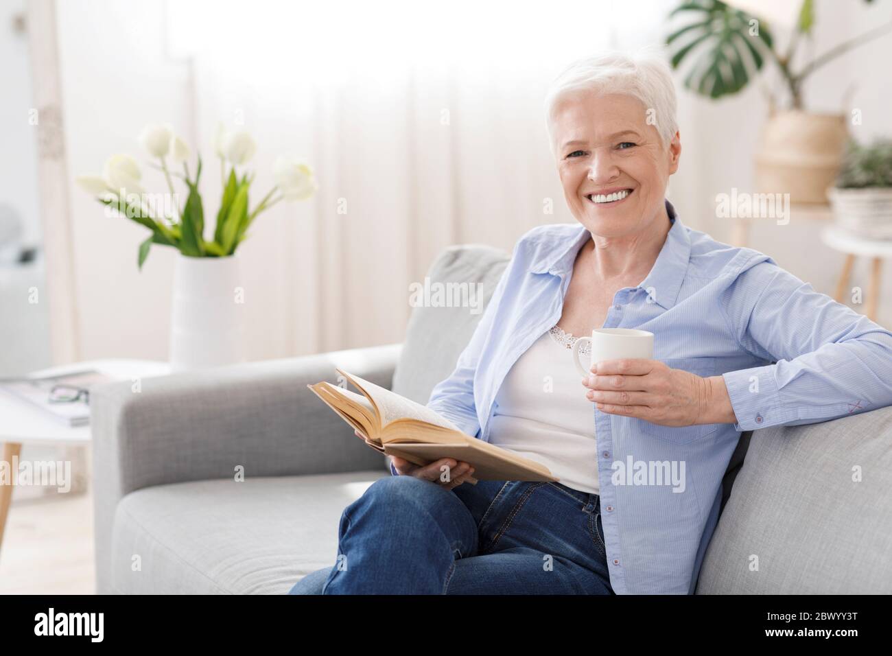Lieblingsbeschäftigung. Ältere Frau, die sich zu Hause mit einem Buch und heißem Tee entspannt Stockfoto