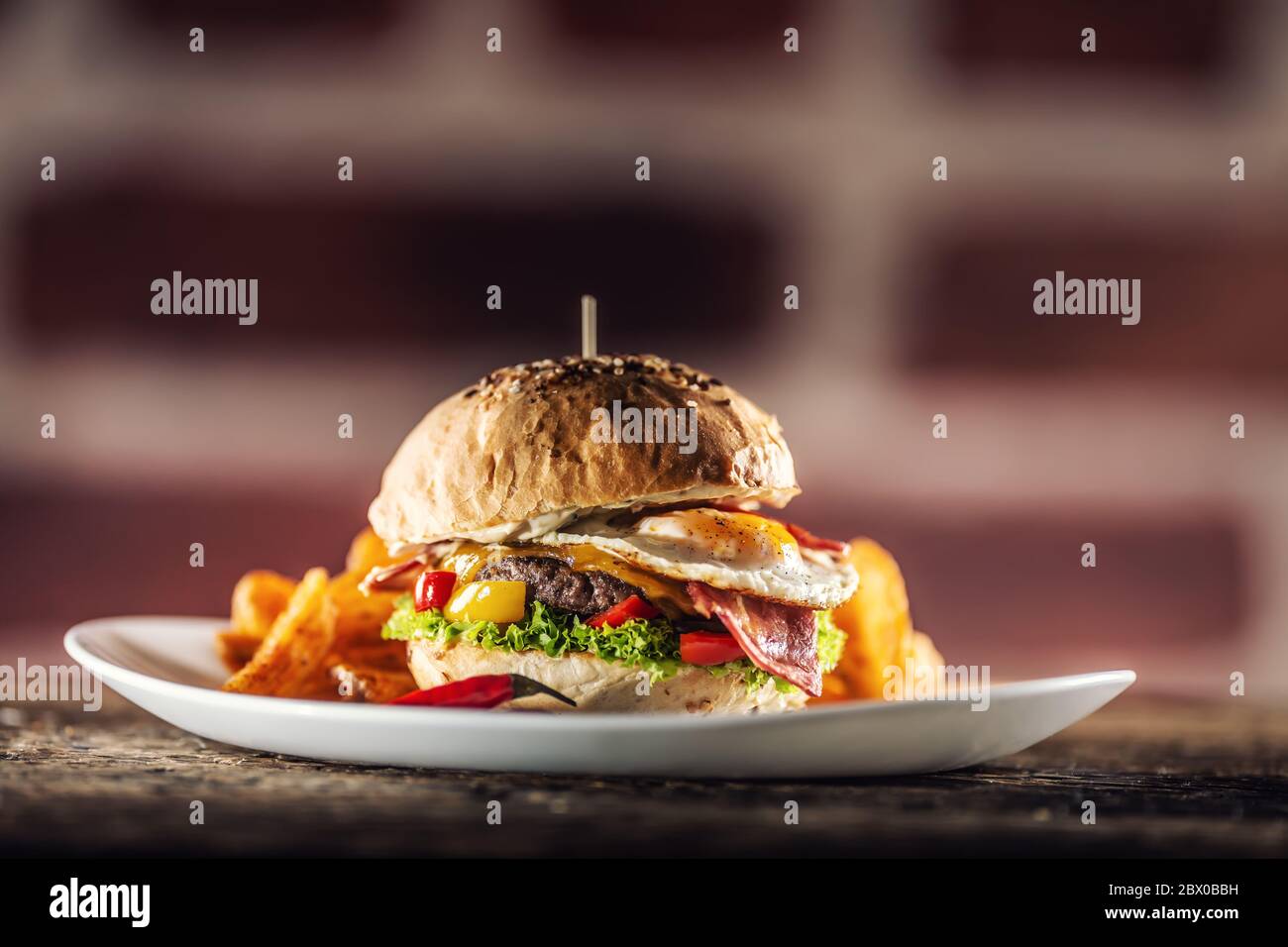Rinderburger mit Spiegelei, Speck, Käse, Salat und Paprika in einem Sesambrötchen mit Kartoffelkeilen mit geräuchertem Paprika und extra frischem Chili auf dem Stockfoto