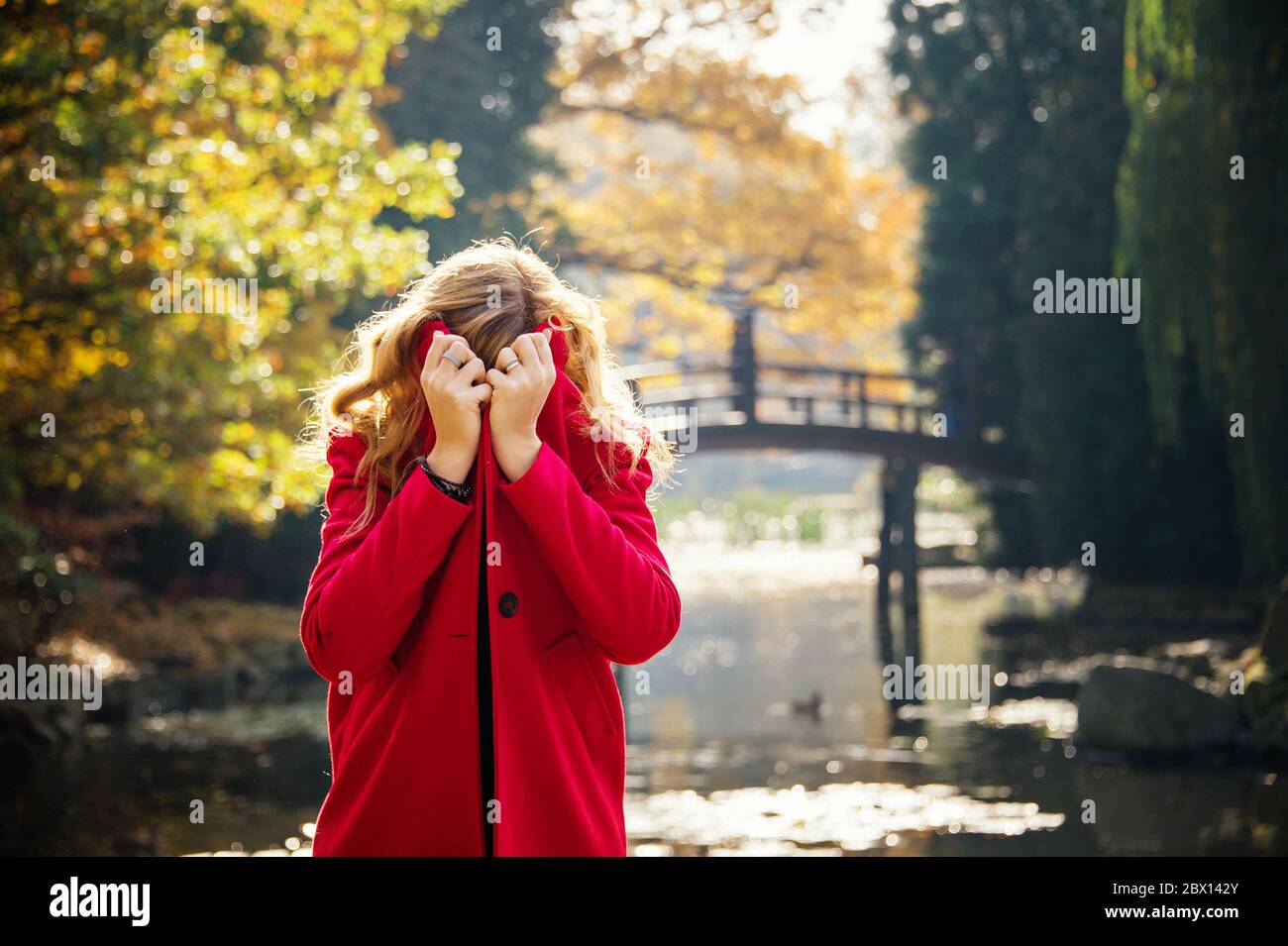Junge Frau versteckt sich unter rotem Mantel im Herbst Stadtpark in der Nähe des Sees Stockfoto