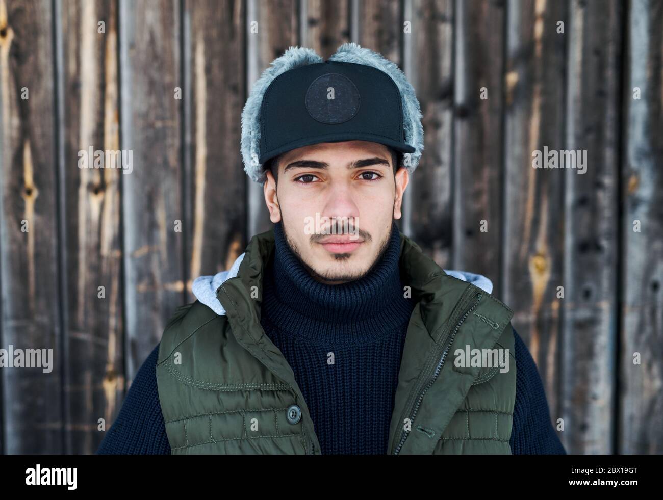 Junger Mann mit Mütze steht im Winter draußen vor Holzhintergrund. Stockfoto