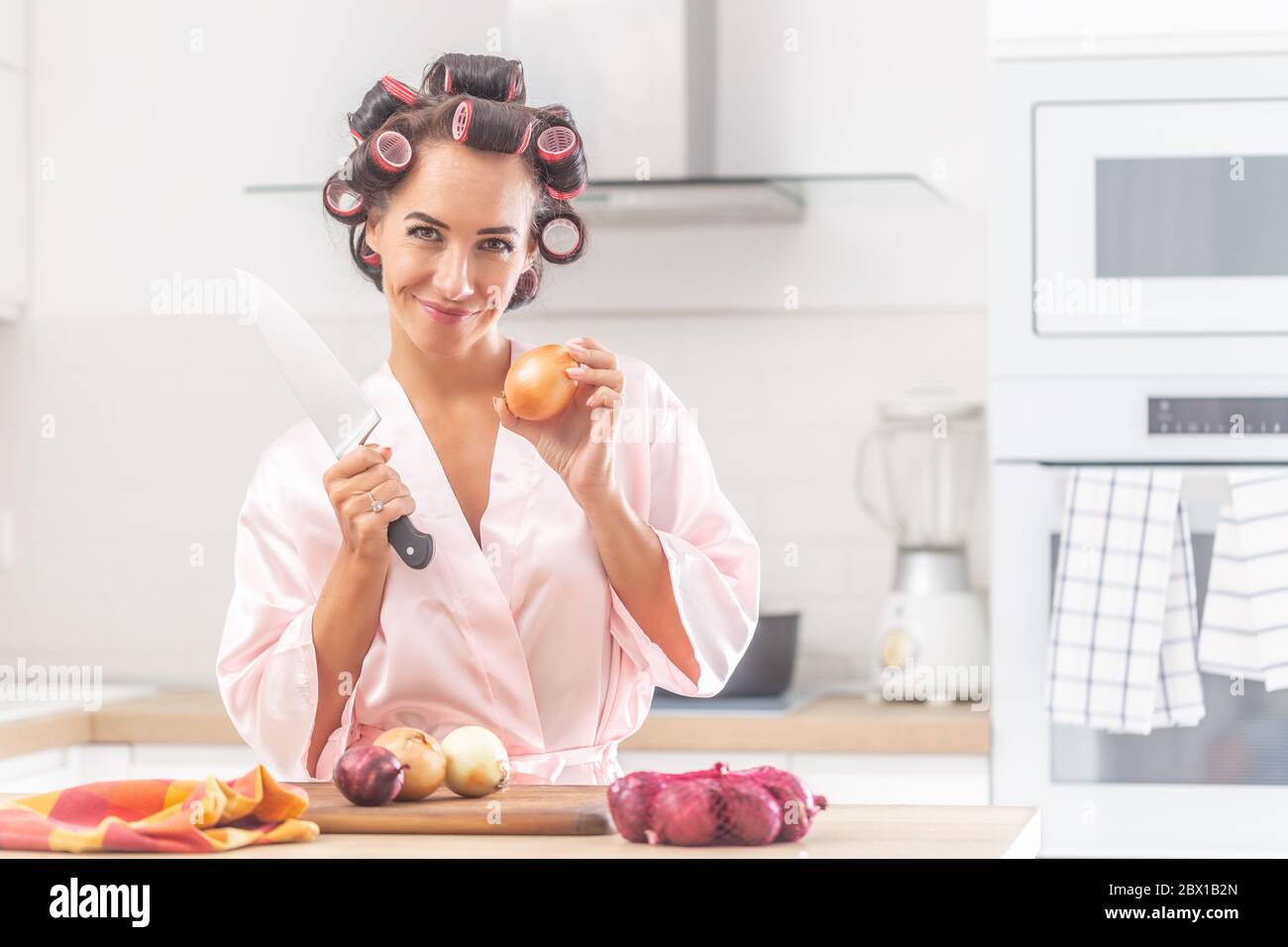 Gut aussehende Frau steht in der Küche hält Messer und eine Zwiebel, tragen Lockenwickler und Nachthemd. Stockfoto