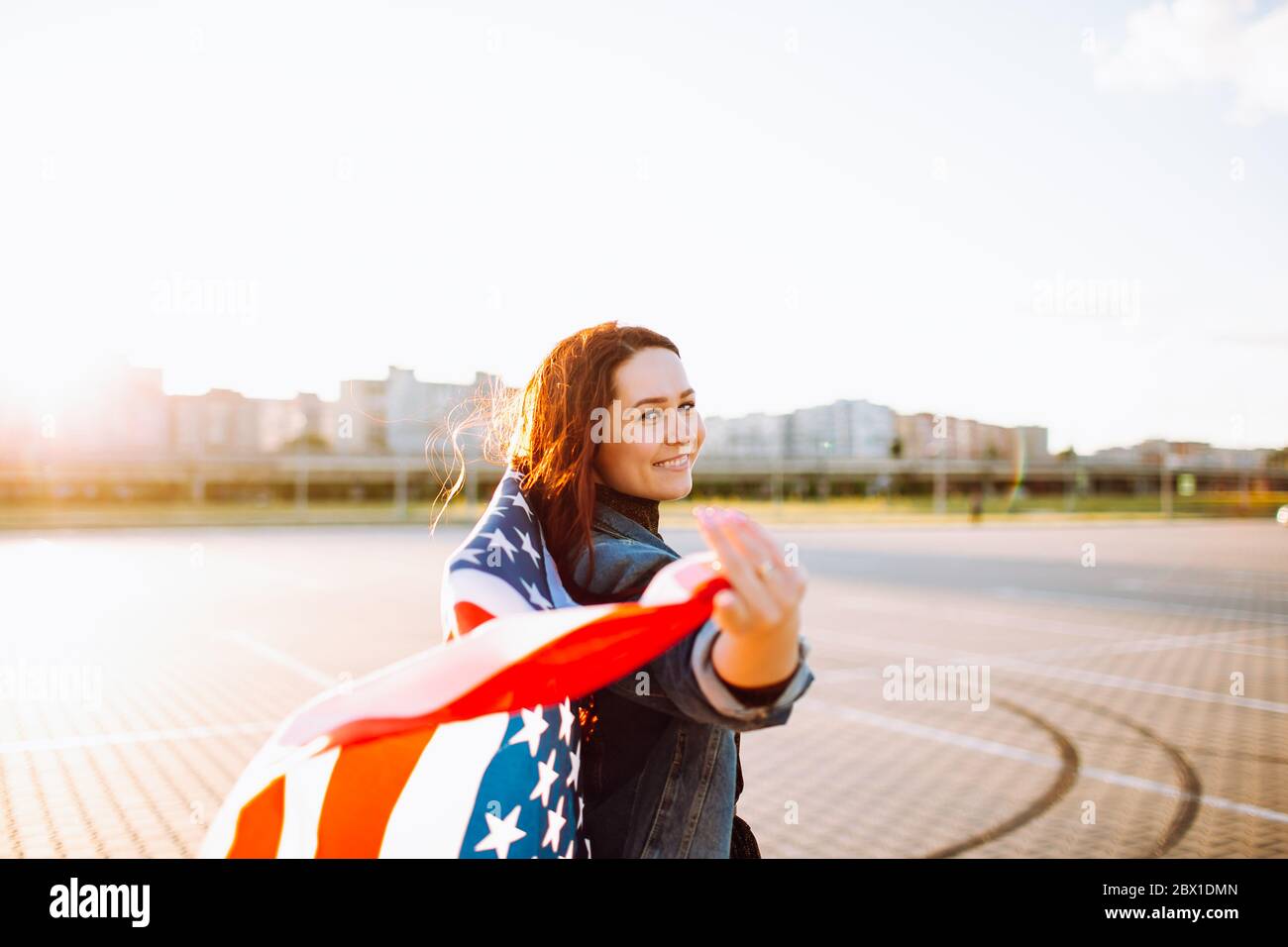 Junge hübsche rothaarige Frau mit nationaler us-Flagge über Sonnenschein gewickelt. Weiche Hintergrundbeleuchtung. Unabhängigkeitstag, amerikanischer Traum, Freiheitskonzept. Stockfoto