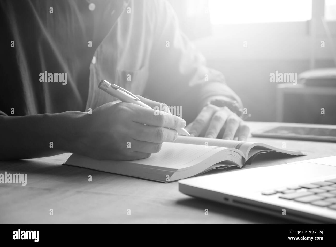Nahaufnahme eines zwanglosen Mannes, der einen Notizbericht schreibt und einen Laptop auf dem Schreibtisch benutzt, der von Sonnenlicht durchstrahlt wird Stockfoto