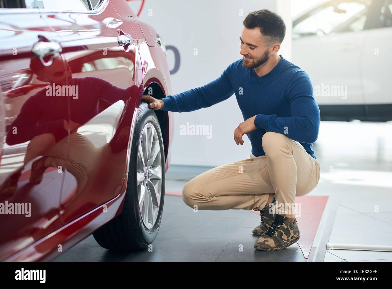 Schöner lächelnder bärtiger Mann, der sich hinsetzt, berührt und auf das Rad des roten Autos im Autohaus schaut. Seitenansicht des glücklichen Nachwuchses vor dem erfolgreichen Kauf. Stockfoto