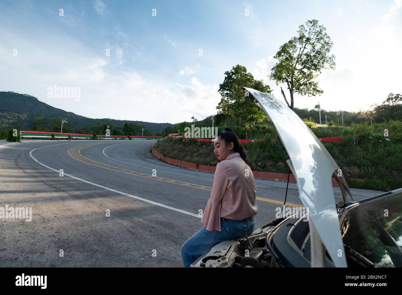 Eine junge schöne Dame sitzt in der Nähe des Autos für die Hilfe auf der öffentlichen Straße im Wald bei Berg-und Himmel Hintergrund zu rufen Stockfoto
