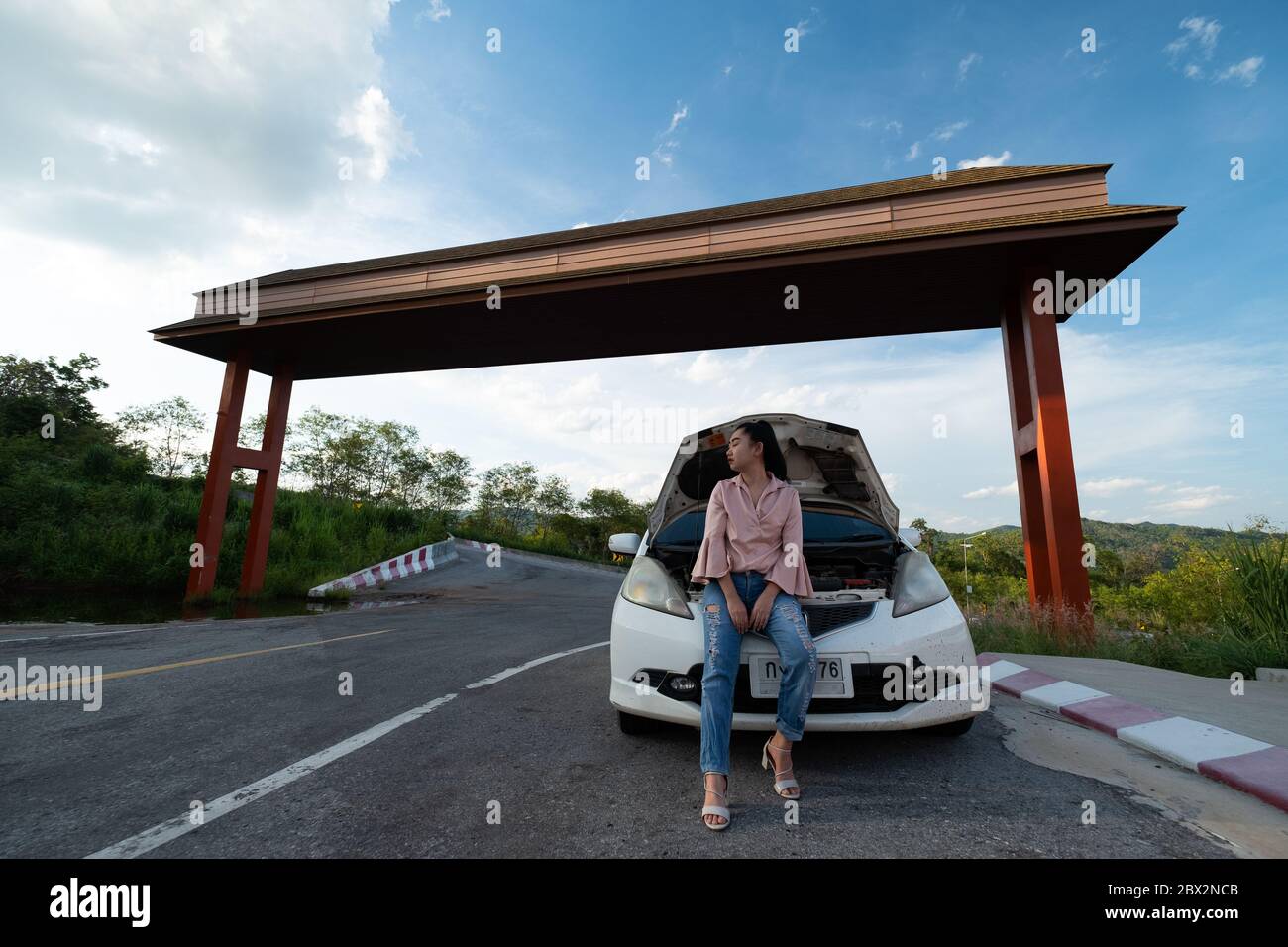 Eine junge schöne Dame sitzt in der Nähe des Autos für die Hilfe auf der öffentlichen Straße im Wald bei Berg-und Himmel Hintergrund zu rufen Stockfoto