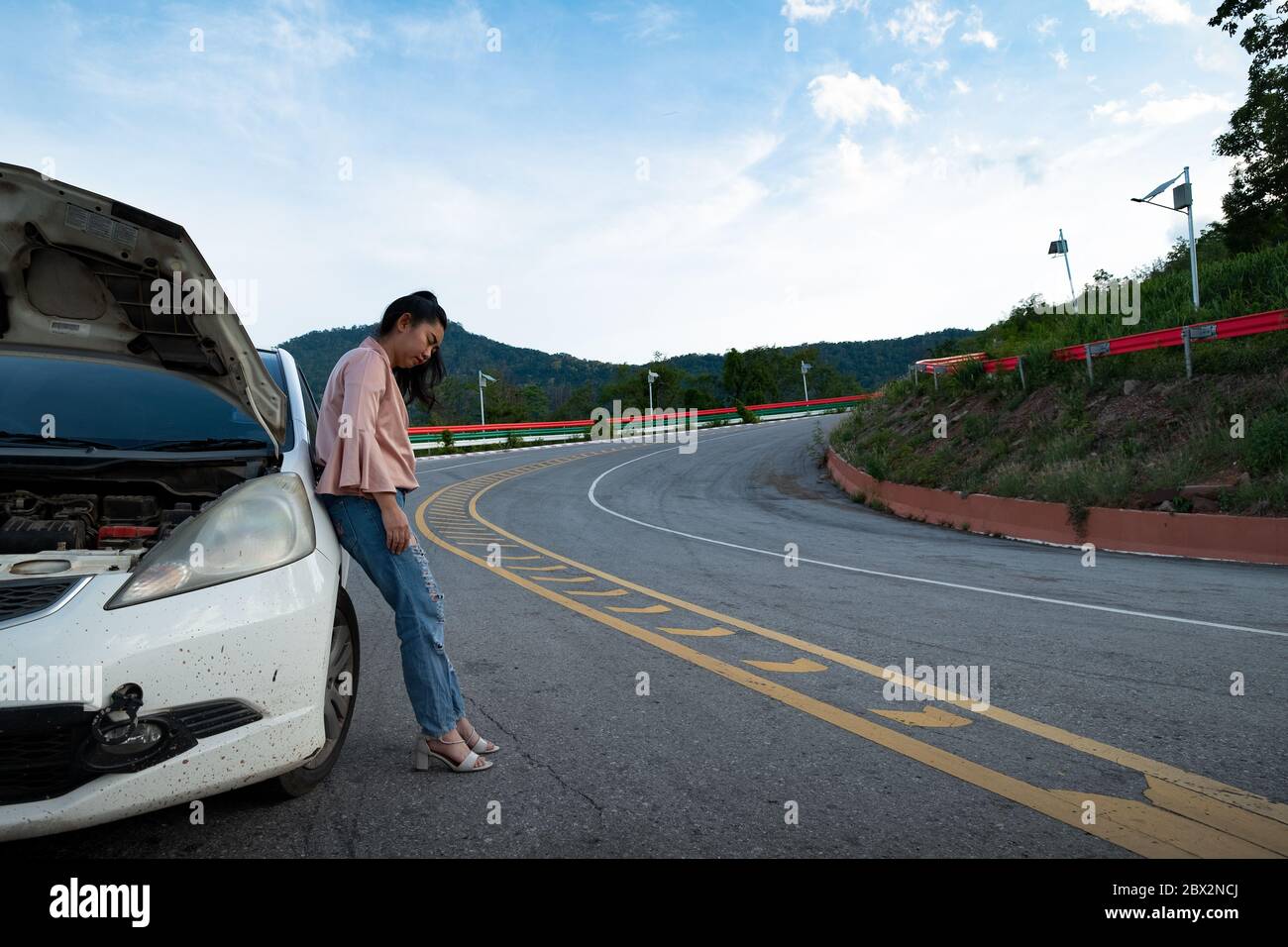 Eine junge schöne Dame steht in der Nähe des Autos für die Hilfe auf der öffentlichen Straße im Wald bei Berg-und Himmel Hintergrund zu rufen Stockfoto
