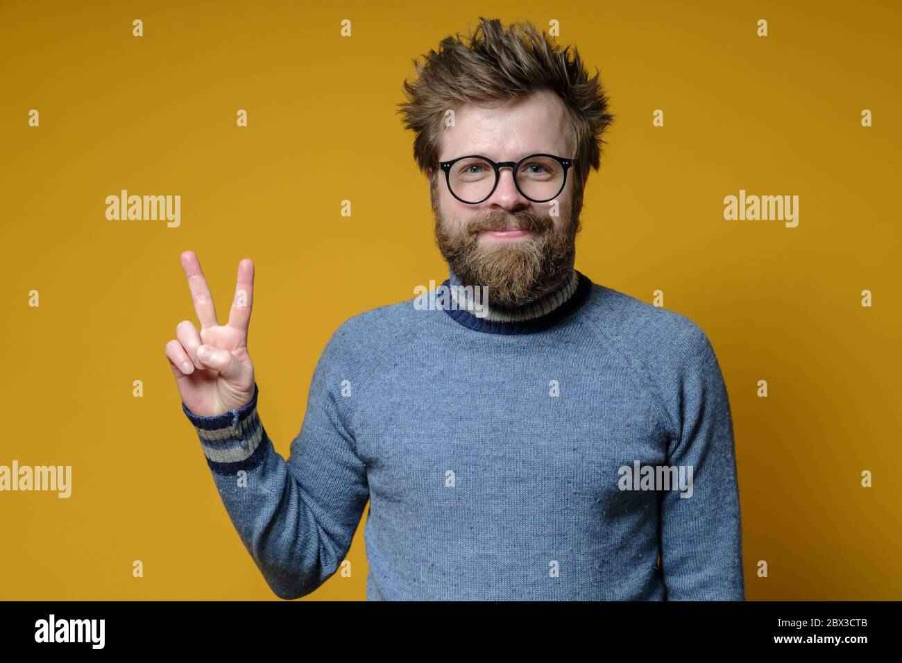 Positiver Mann in der Brille macht ein V-Zeichen mit den Fingern. Der zottige, bärtige Hipster in einem alten Pullover macht mit der Hand eine Siegesgeste. Stockfoto