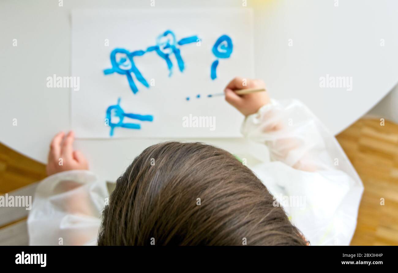 Kleiner Junge, der mit einem Pinsel und blauer Farbe und einem weißen Papier auf einem weißen Tisch malt Stockfoto