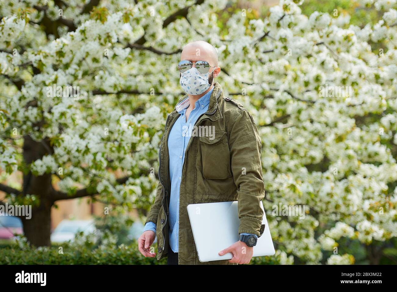 Ein Glatzkopf mit Bart in einer medizinischen Gesichtsmaske, um die Ausbreitung des Coronavirus zu vermeiden, geht mit einem Laptop in den Park. Ein Mann trägt eine n95 Gesichtsmaske und einen Piloten Stockfoto