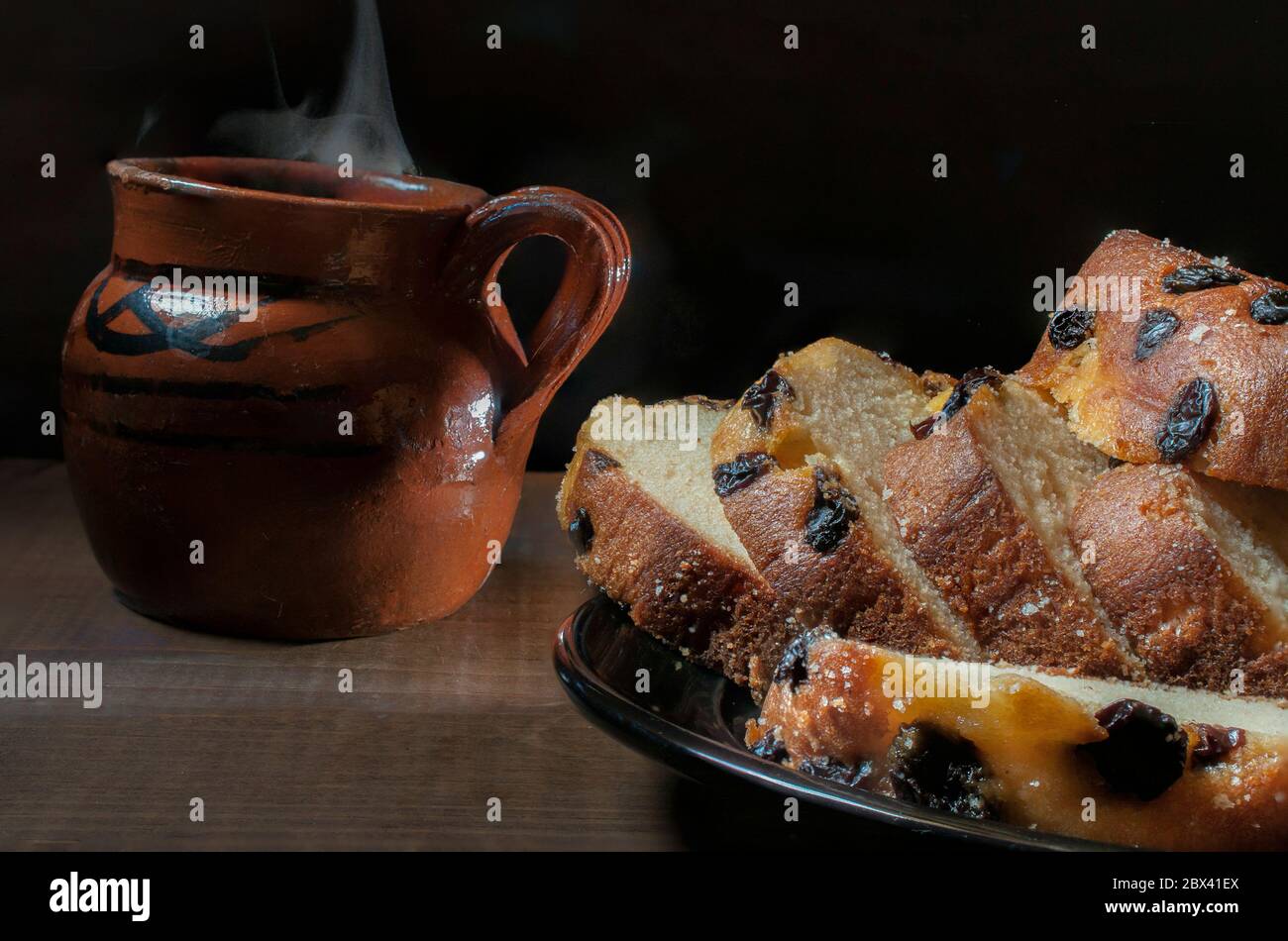 Kuchenscheiben mit Rosinen auf einem schwarzen Teller und einem mexikanischen Ton- oder Schlamm-Kaffeebecher, auf dem Hintergrund auf einem rustikalen Tisch und in einer dunklen Umgebung. Stockfoto