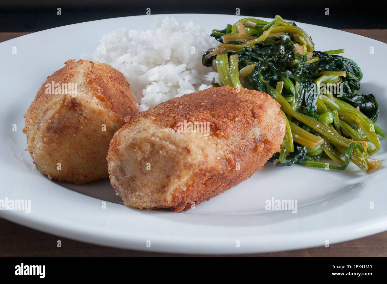 Nahaufnahme von Thunfischkroketten mit Dampfreis und Spinat auf einem weißen Teller in einem dunklen Hintergrund Stockfoto