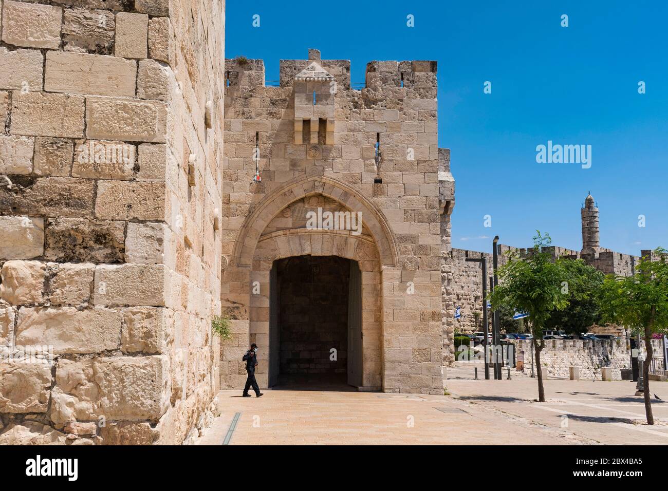 Ein einjähriger maskierter Polizist, der während der Quarantäne am jaffa-Tor der Altstadt jerusalems mit dem Turm davids im Hintergrund vorbeigeht Stockfoto