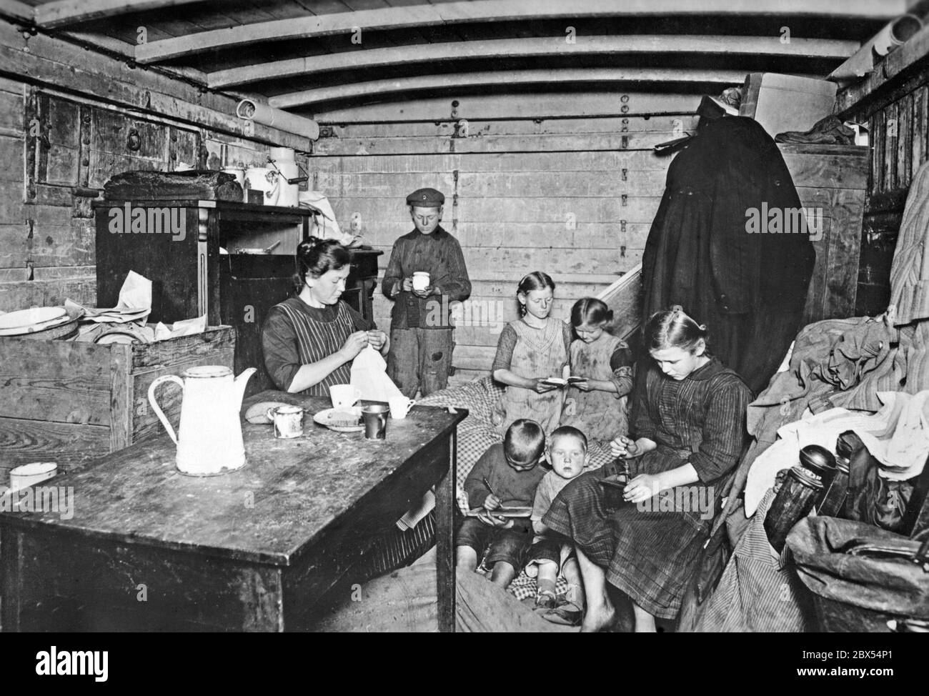 Die Mutter und sechs Kinder sitzen in einem Güterwagen, der in eine Unterkunft umgewandelt wurde. Die Mutter saut, die Kinder lesen oder schreiben auf einen Schiefertafel. Stockfoto