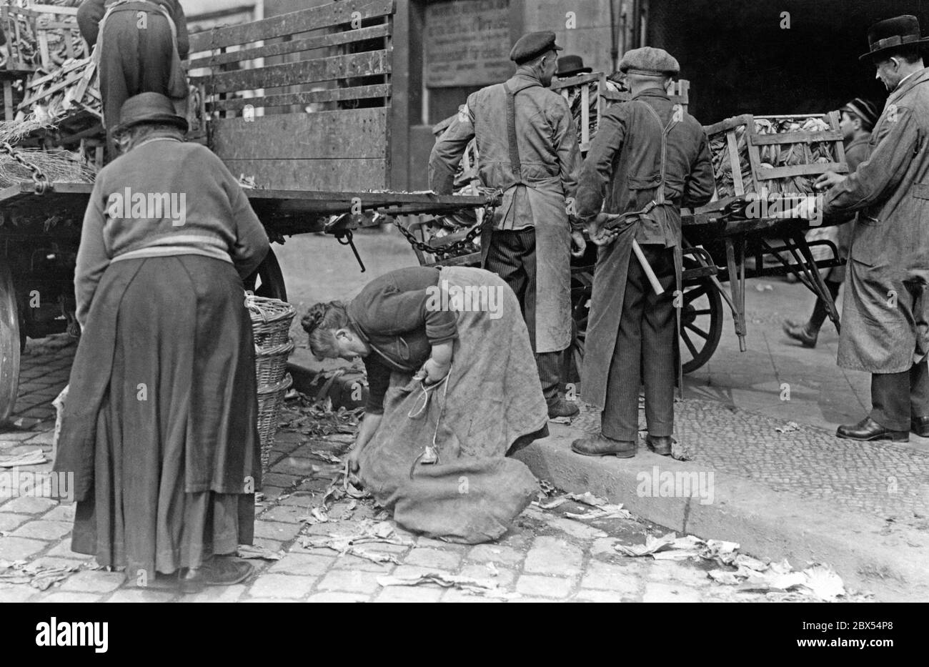 Zwei Frauen sammeln die Lebensmittelabfälle aus der Großmarkthalle. Händler transportieren neu gelieferte Waren in großen Kisten von einem Wagen zur Markthalle. Stockfoto