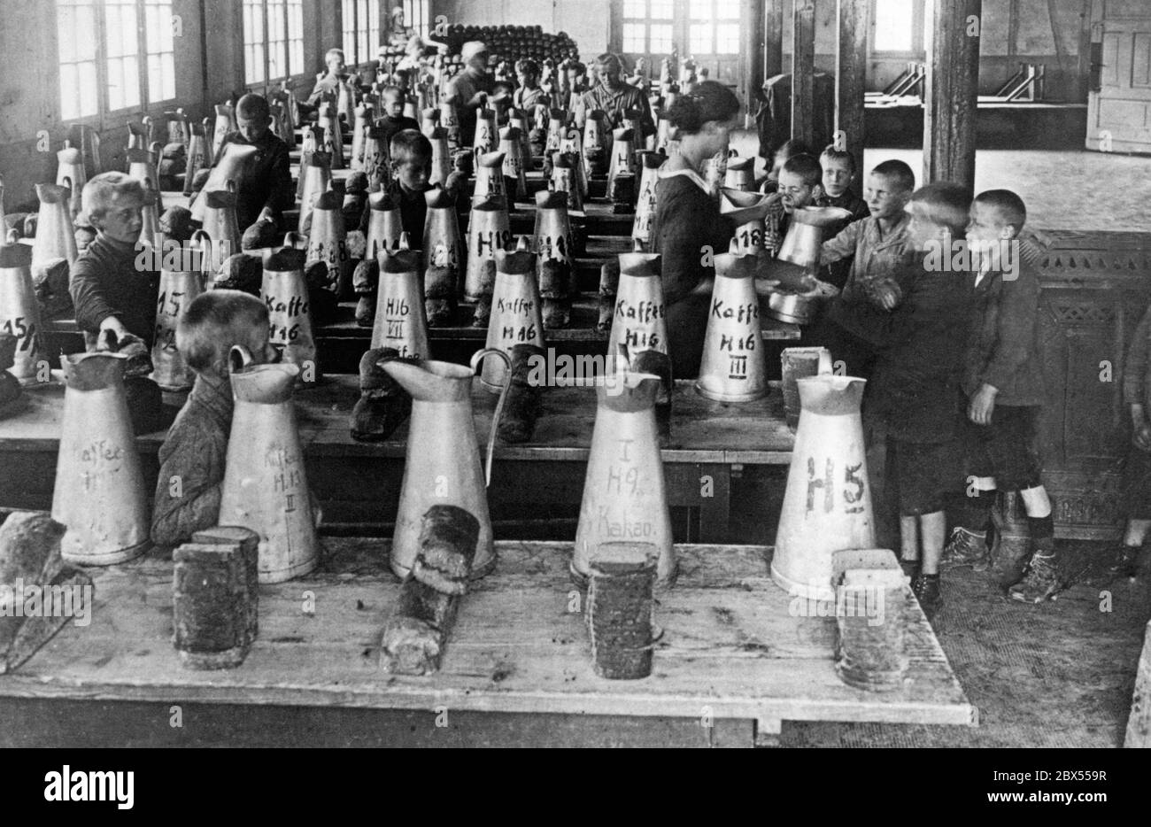 Einige schlecht bekleidete Kinder bekommen Essen und Trinken in einem kargen Zimmer. Auf langen Holztischen gibt es Krüge von Kaffee und Kakao, und vor ihnen sind Stapel von Brot in Scheiben. Auf dem Höhepunkt der Inflation sind in diesem süddeutschen Erholungslager 3000 Stadtkinder aus armen Quartieren untergebracht, weil ihre Eltern sie nicht mehr ernähren können. Um 1923. Stockfoto