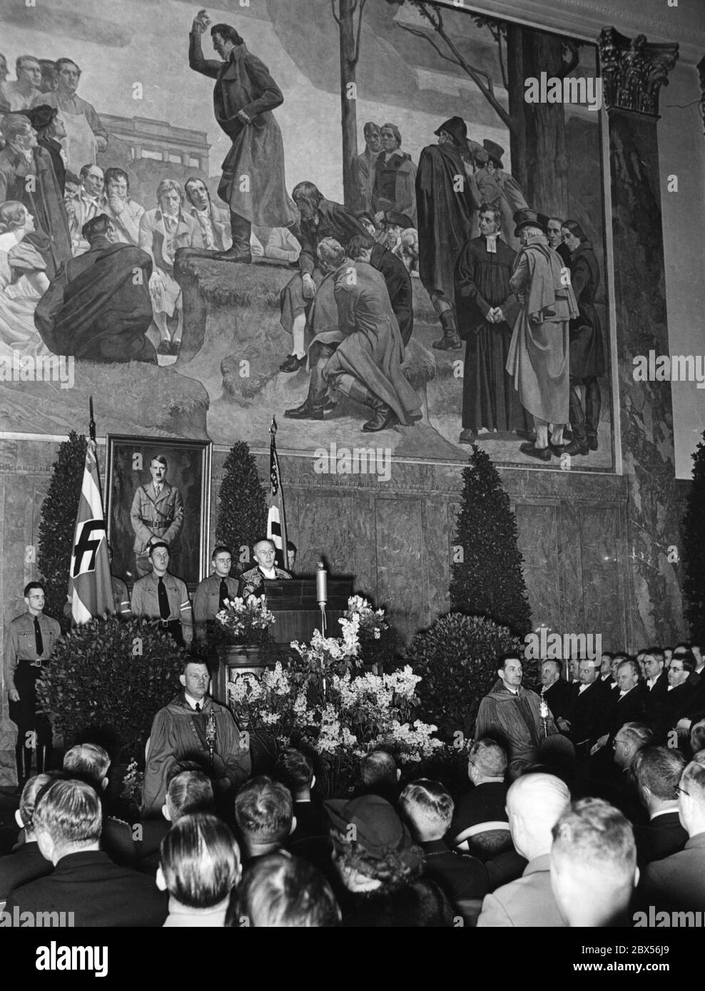 Der Rektor der Universität Berlin, Professor Willy Hoppe (am Pult in Kleid), begrüßt die Anwesenden bei einer Feier zum 5. Jahrestag des "Tags der nationalen Erhebung" in der Neuen Aula. Hinter ihm steht ein Porträt Adolf Hitlers. Stockfoto