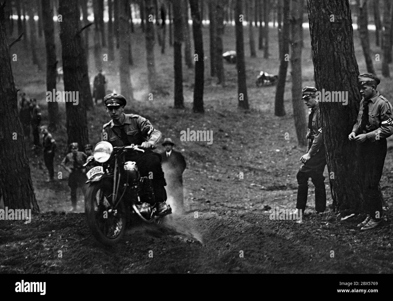 Ein Mitglied der Motor HJ fährt mit seinem DKW SB 200A Motorrad durch den Wald bei einer Offroad-Fahrt. Andere Mitglieder der Hitlerjugend stehen am Rande der Strecke. Stockfoto